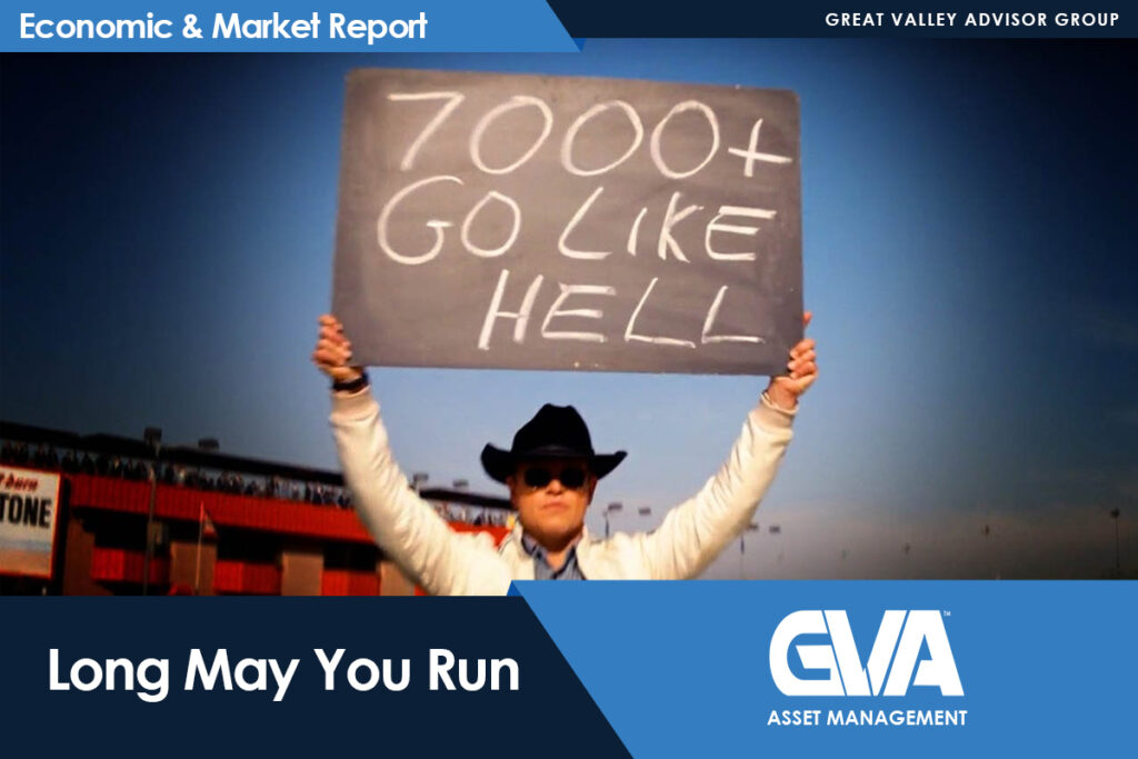 Person in a hat and sunglasses holds up a sign reading '7000+ GO LIKE HELL' at a stadium, with 'Economic & Market Report' branding visible above.